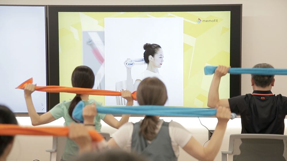 스크린 영상을 따라 탄력밴드를 들어 올리는 단체 운동교실
