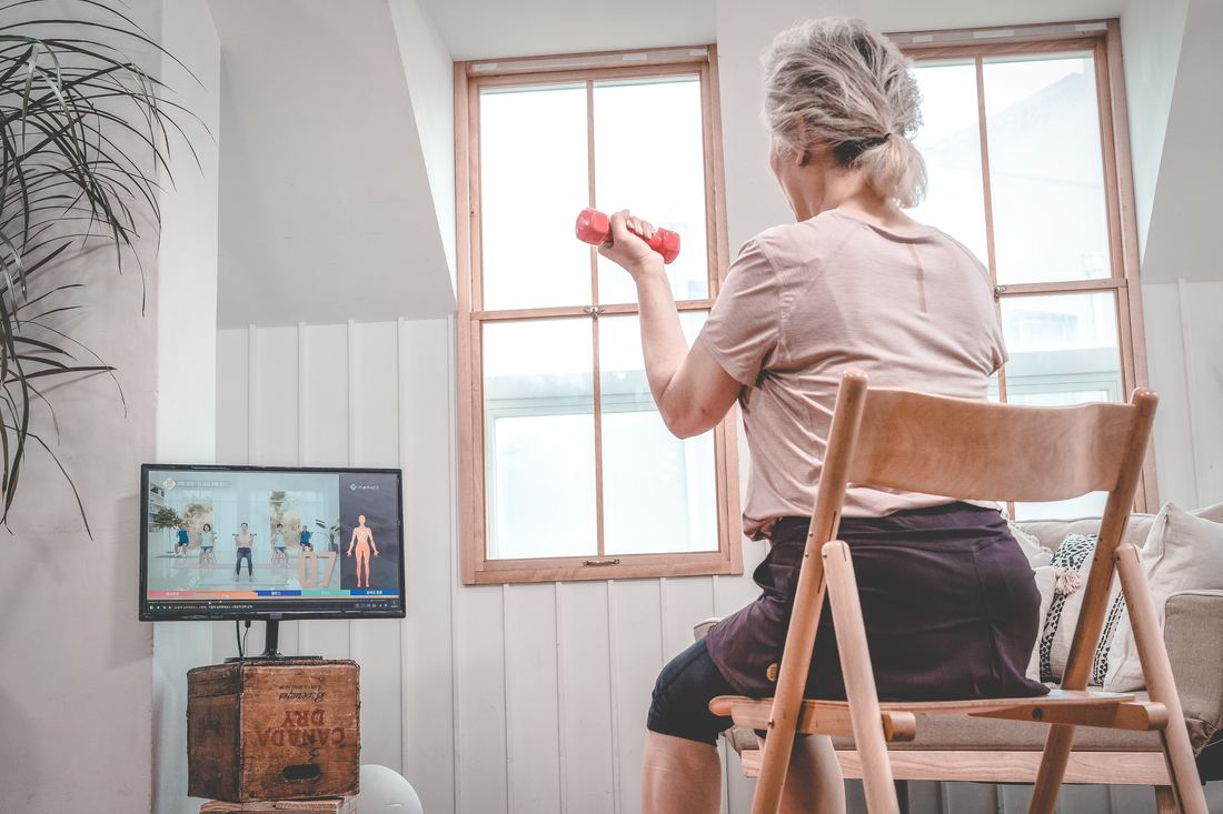집에서 TV 운동 영상을 보며 의자에 앉아 아령 운동을 하는 시니어